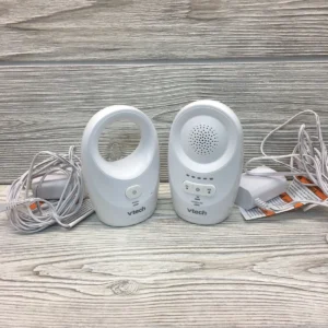 VTech DM1111 Digital Audio Baby Monitor a white VTech audio baby monitor set, including a parent unit (left), a baby unit (right), and two power adapters with UK-style plugs, all resting on a patterned textile surface.