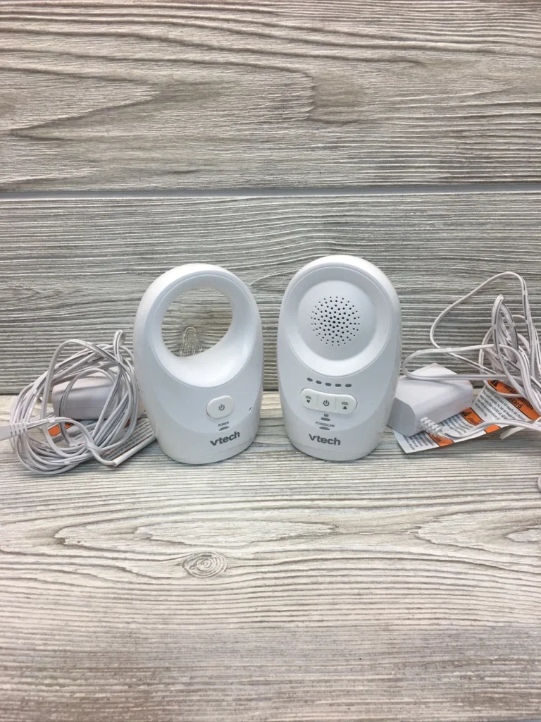 VTech DM1111 Digital Audio Baby Monitor a white VTech audio baby monitor set, including a parent unit (left), a baby unit (right), and two power adapters with UK-style plugs, all resting on a patterned textile surface.